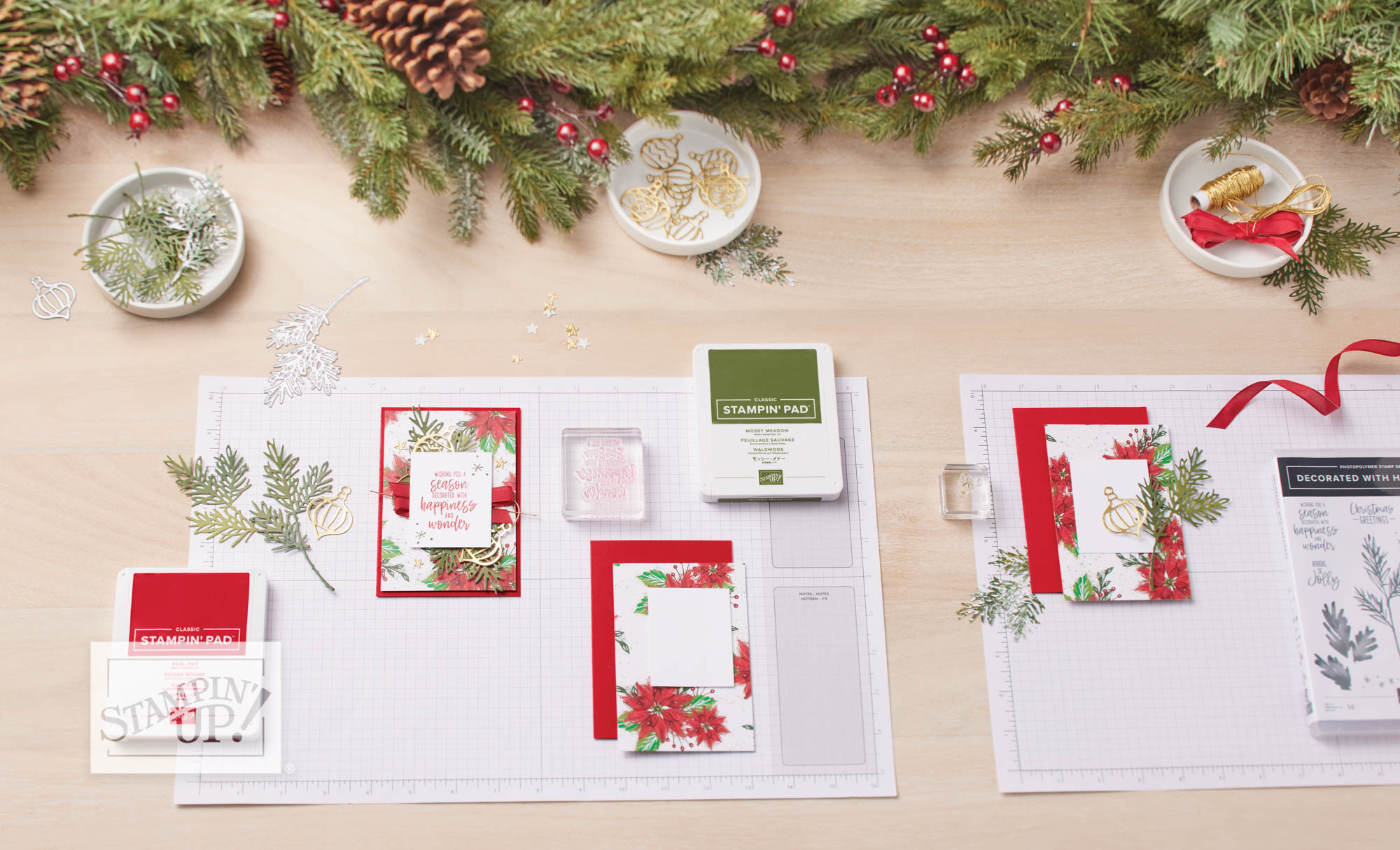 Christmas-themed cardmaking setup featuring pine sprigs, red ribbon, gold embellishments, and festive Stampin’ Up! supplies. Two finished holiday cards are displayed on a grid mat, with coordinating stamps, ink pads, and die cuts laid out for crafting.