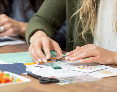 Close-up of a woman using a clear acrylic block to stamp an image onto cardstock during a cardmaking session. A crafting table is filled with paper, stamps, scissors, and embellishments, with another crafter visible in the background.